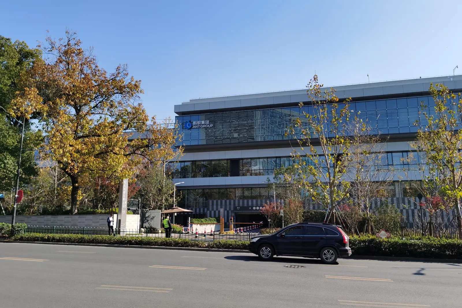 A modern office building with an Ant Group sign behind autumn trees and a street in Hangzhou on a sunny day.