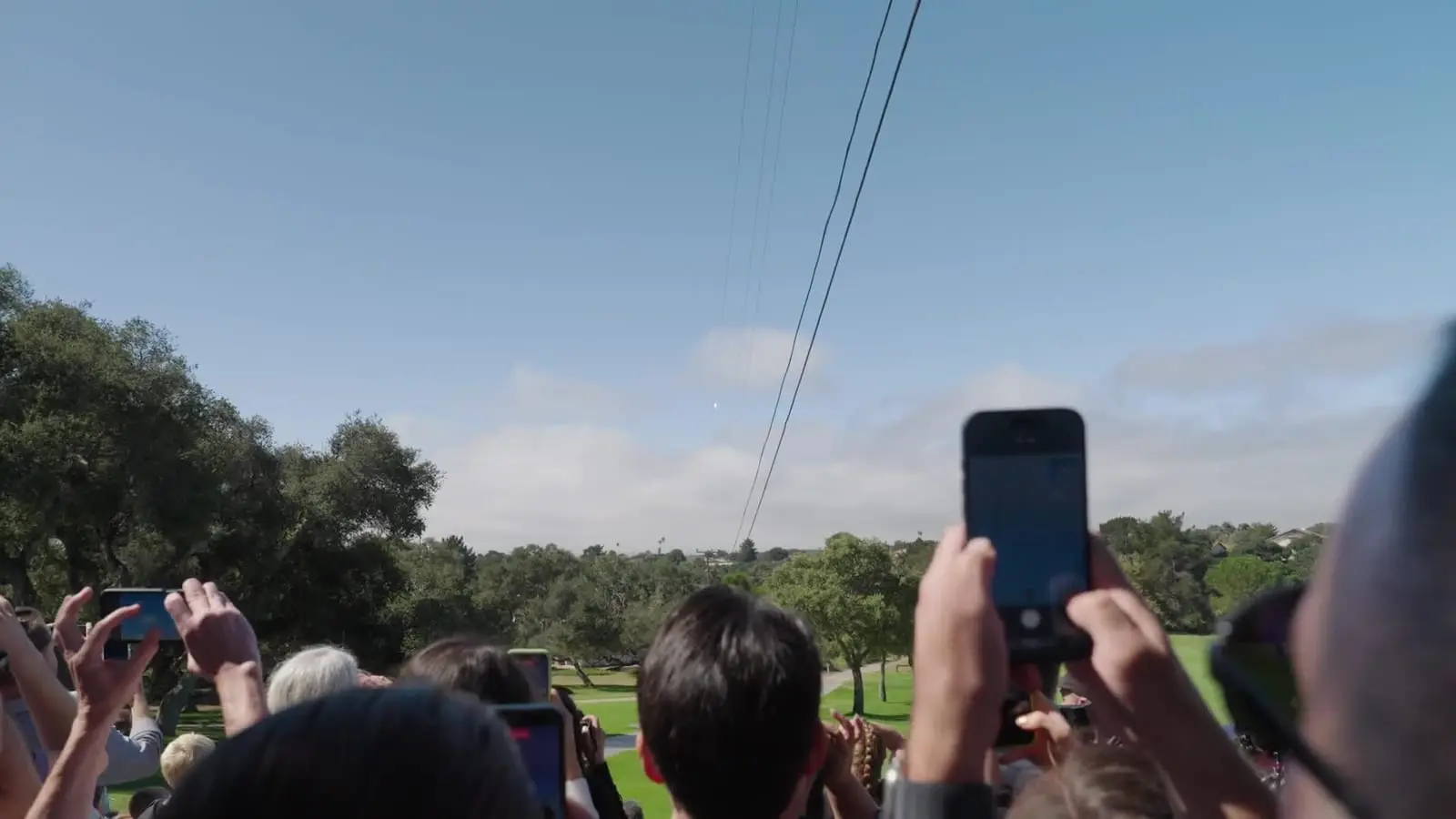 A crowd under a blue sky raises phones to film a rocket climbing in the distance above trees.