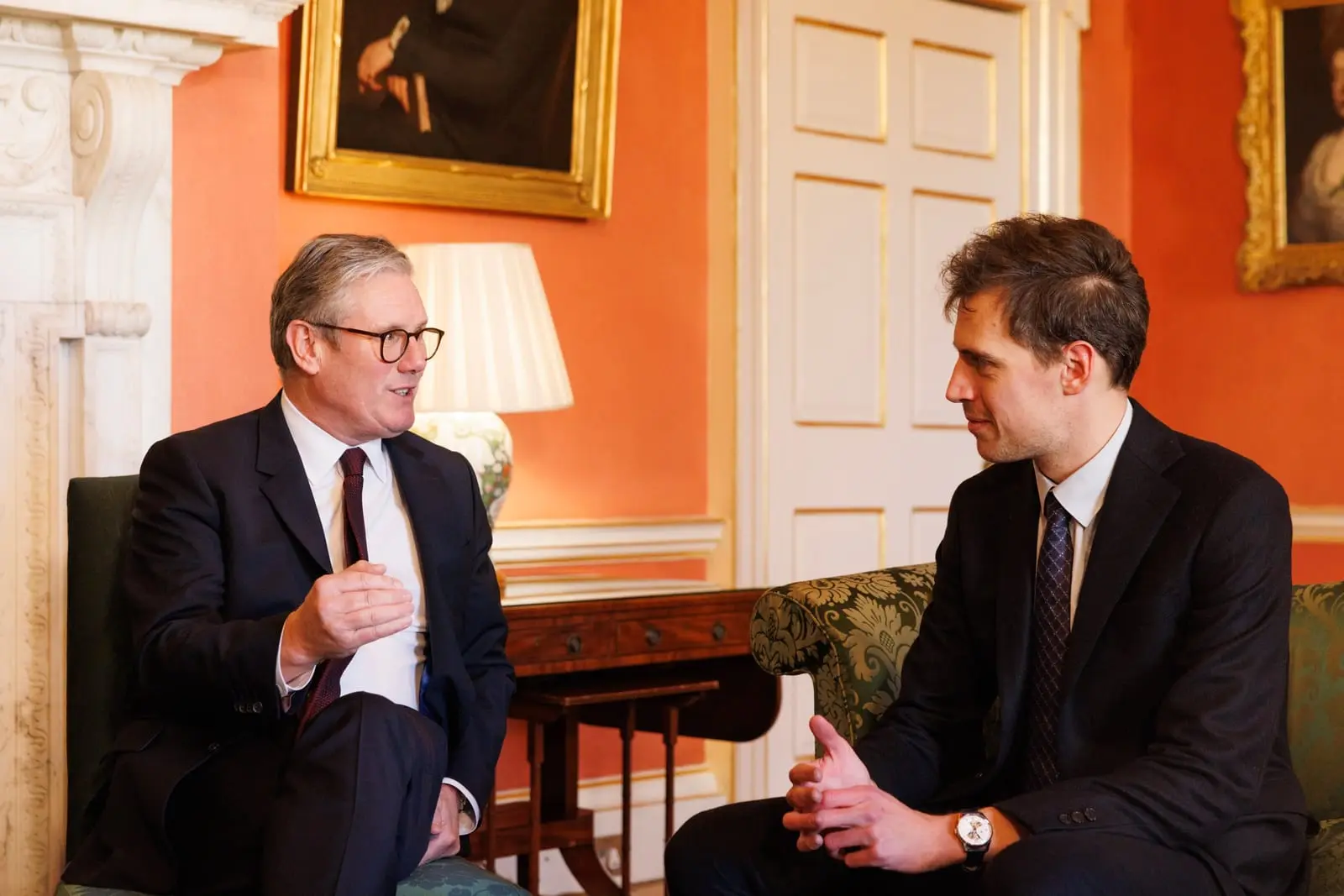 Two men in dark suits sit facing each other in armchairs and talk in an orange-walled room with framed portraits.