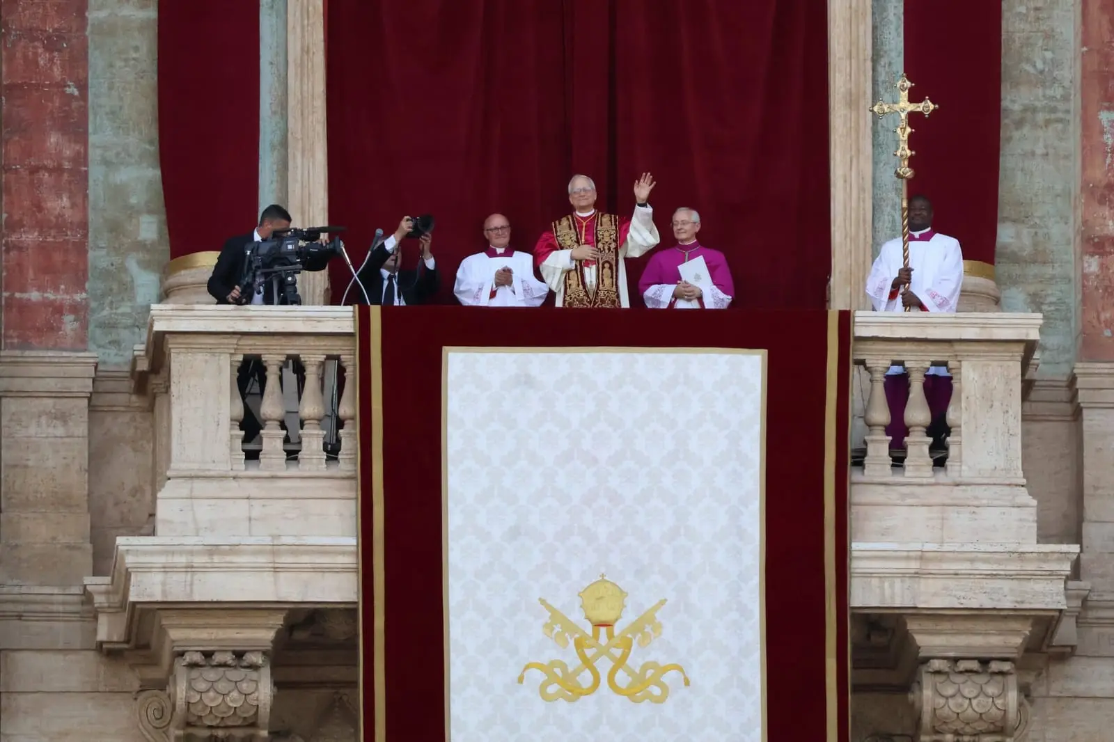 Pope Leo XIV in red and gold vestments waves from the central balcony of St. Peter's Basilica beside clergy and camera operators.