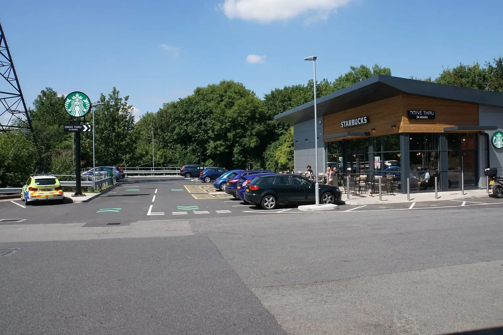 A Starbucks drive-thru building with a wooden facade and a Drive Thru 24 hours sign, with cars parked outside on a sunny day.