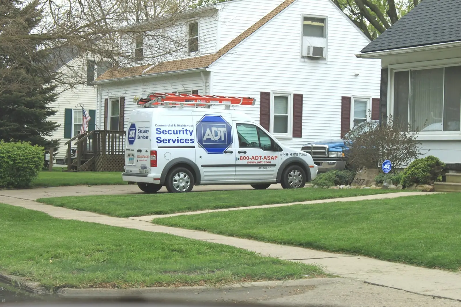 A white ADT Security Services van with a ladder on the roof parked in front of a white house with a green lawn.