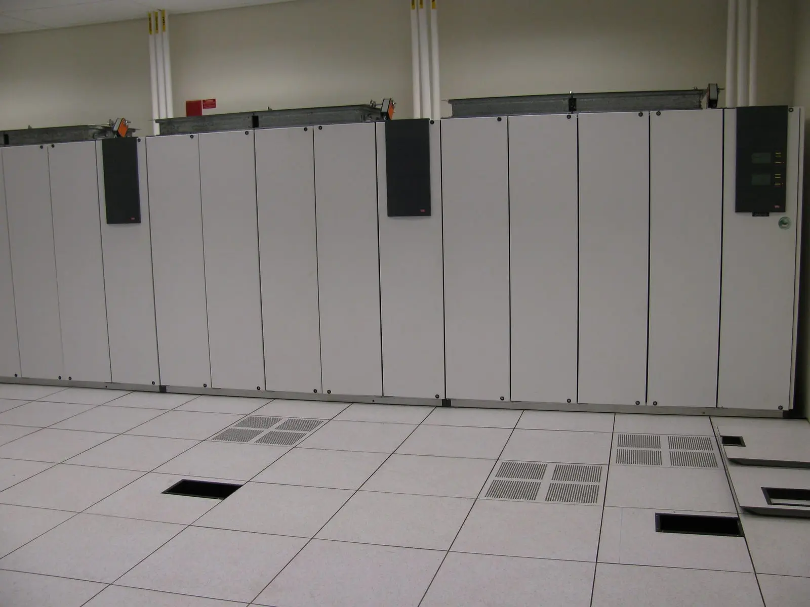 A row of white computer room air conditioning cabinets with dark control panels standing on a raised data center floor with perforated tiles.